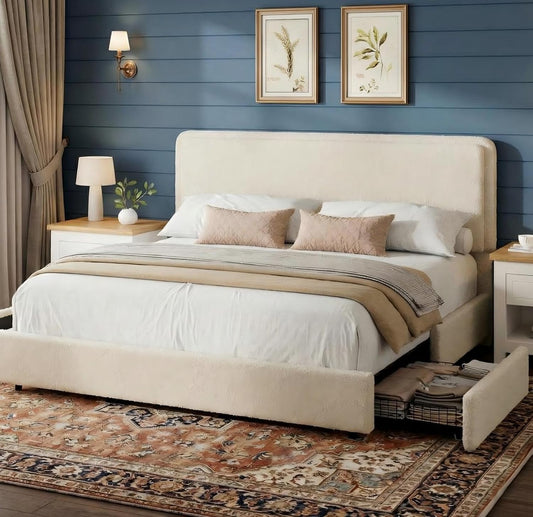 A cream upholstered bed frame with a tall headboard and low footboard, shown in a styled bedroom with white bedding, beige pillows, a patterned rug, blue shiplap wall, and bedside tables.