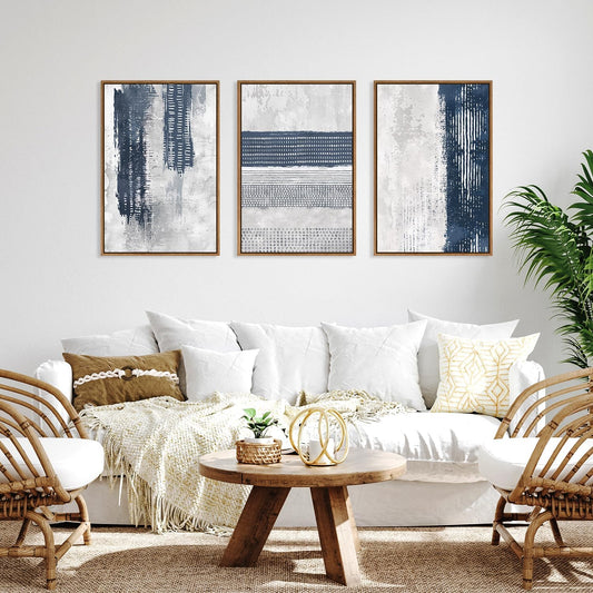 Three framed abstract wall art prints in navy, gray, and white hang above a white sofa in a bright living room with woven chairs, a round wooden coffee table, and a potted plant.