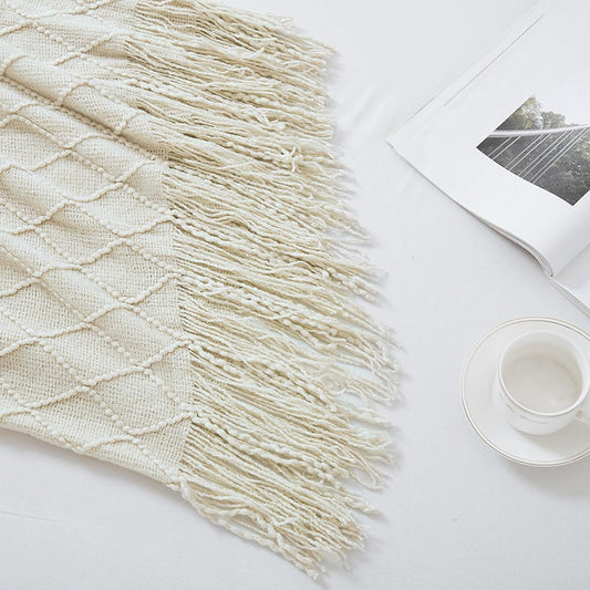 Beige woven blanket with fringe on a white surface with a magazine and cup.