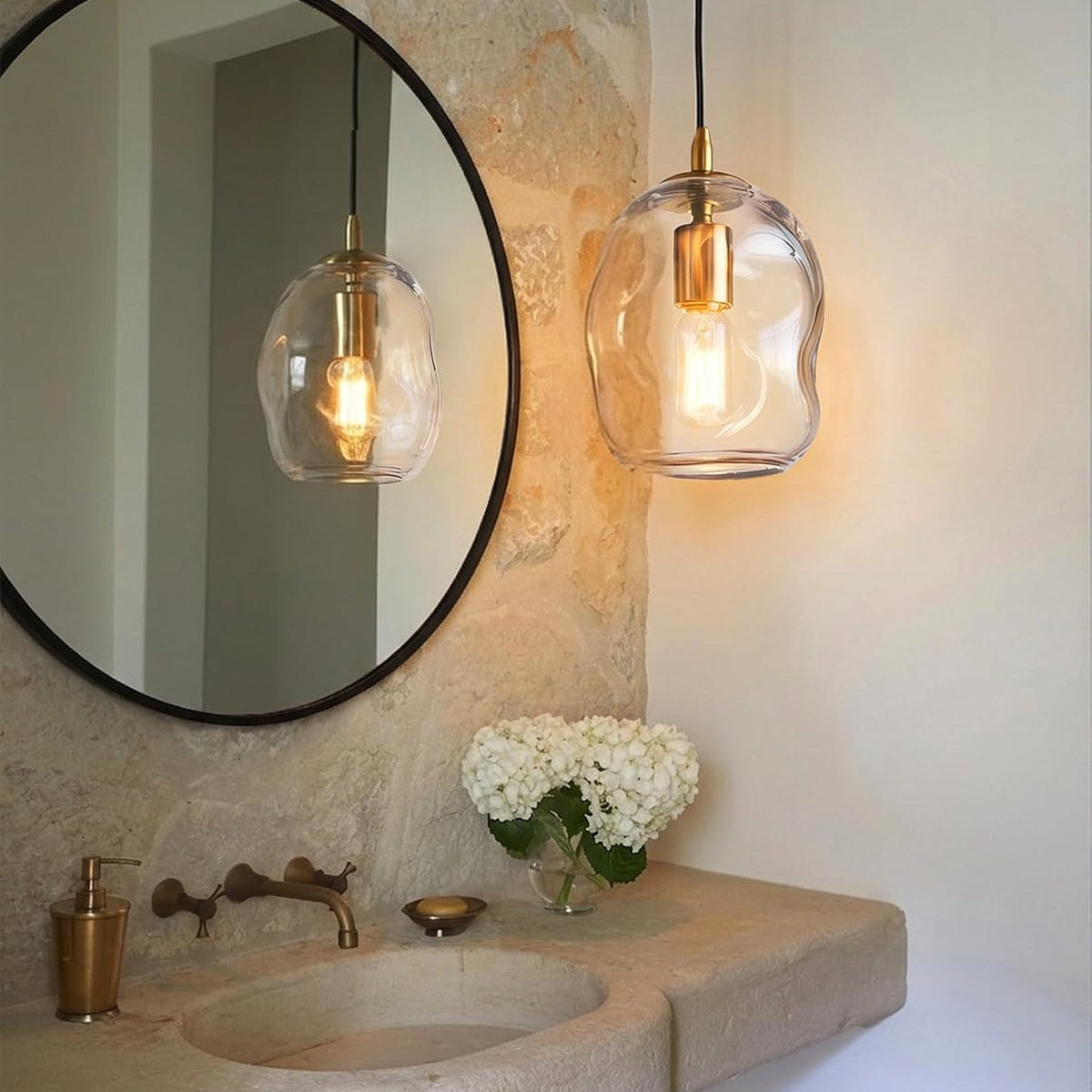 A modern pendant light with an organic, wavy clear glass shade and a brass-colored bulb holder hangs over a stone bathroom vanity next to a large round mirror.