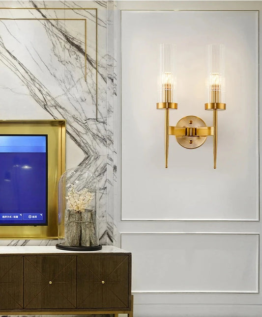 A modern double head gold wall sconce with clear glass lampshades, featuring a gold finish, mounted on a wall.
