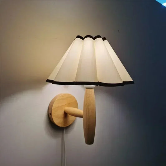 A wall-mounted lamp with a pleated umbrella-shaped shade and a wooden base, illuminated with an LED bulb.