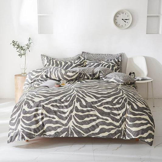 A bed dressed in a zebra-print bedding set with black, gray, and off-white stripes, styled in a bright, modern bedroom with a white chair, side table, and wall clock.