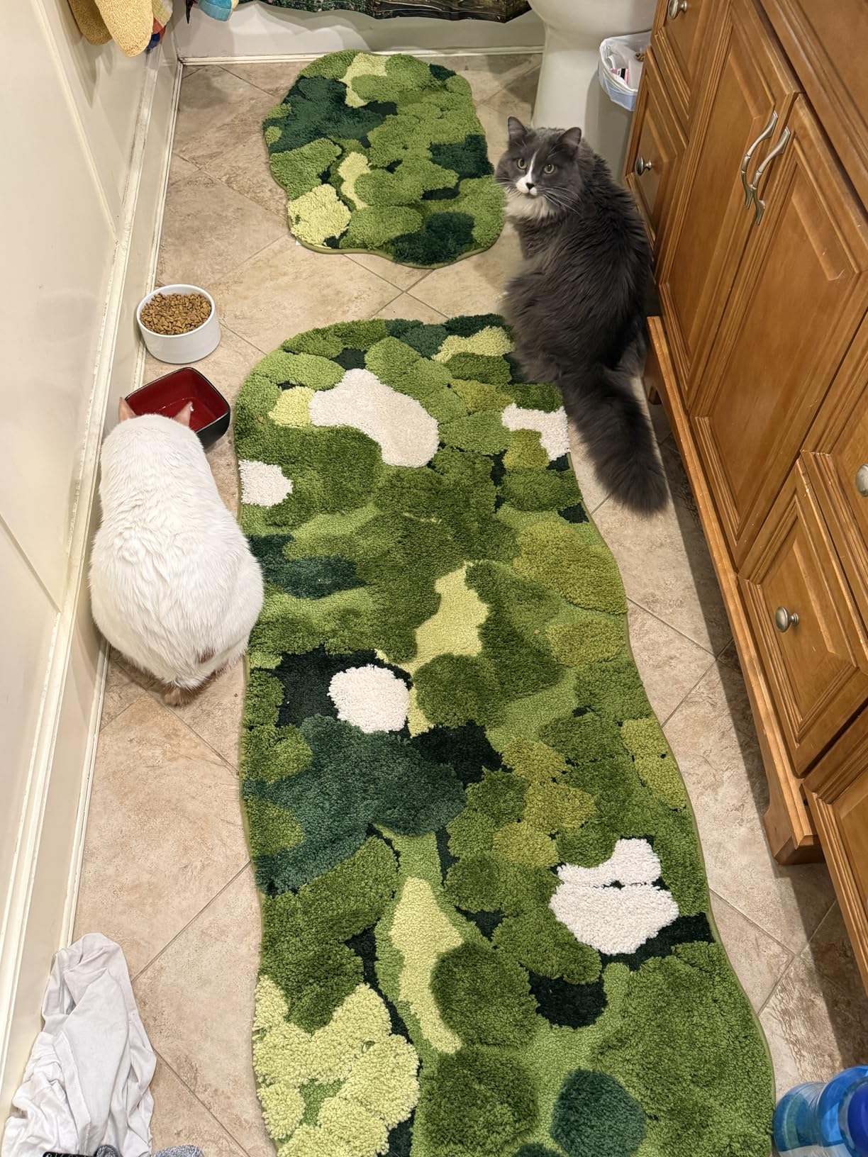 Cat sitting on a green camouflage rug in a bathroom with wooden cabinets and a tiled floor.