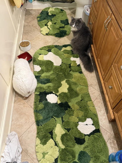 Cat sitting on a green camouflage rug in a bathroom with wooden cabinets and a tiled floor.