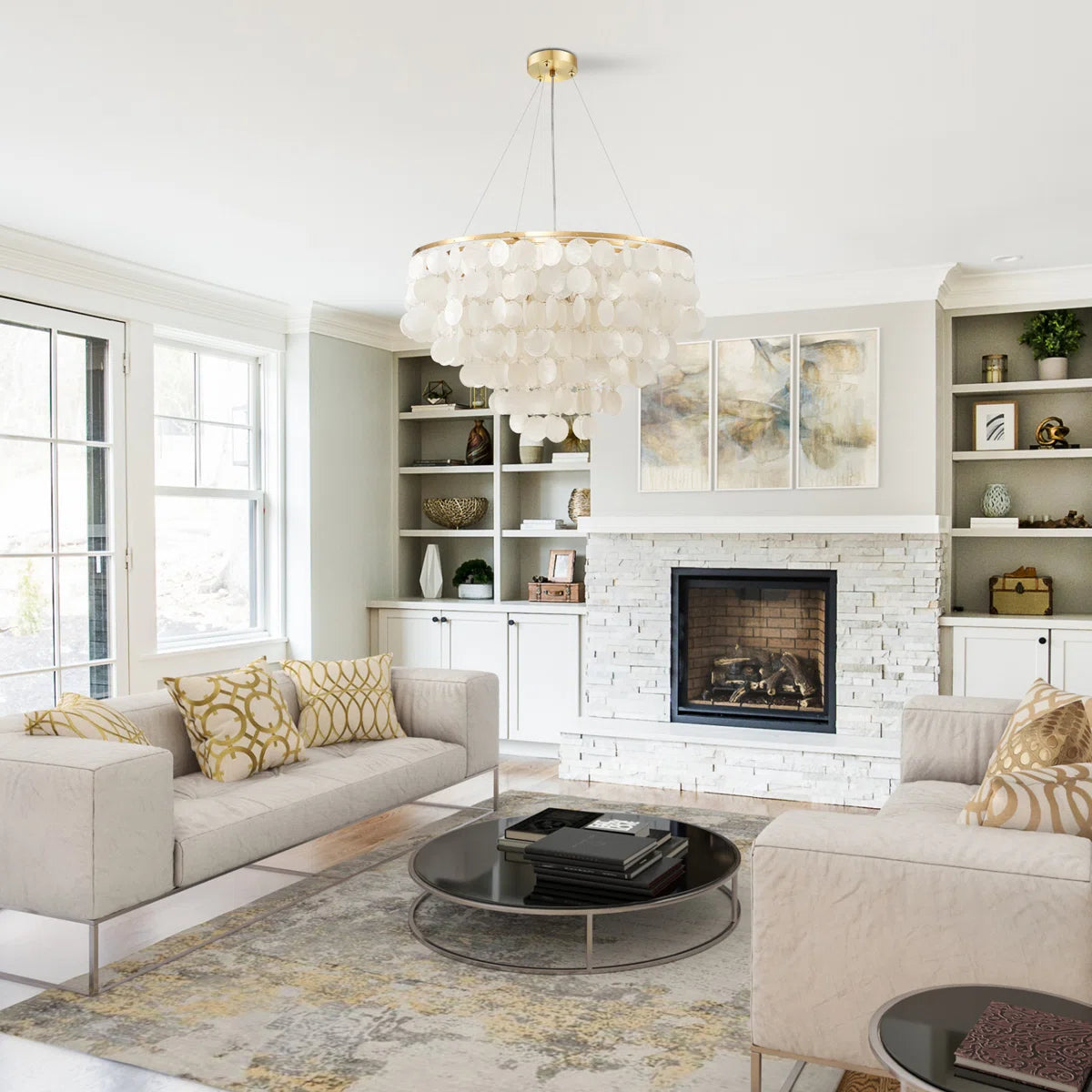A bright, modern living room featuring a large white shell chandelier hanging above a round glass coffee table. The room is styled with a light beige sofa, matching armchair, patterned yellow and white pillows, a white brick fireplace, and built-in white shelves.