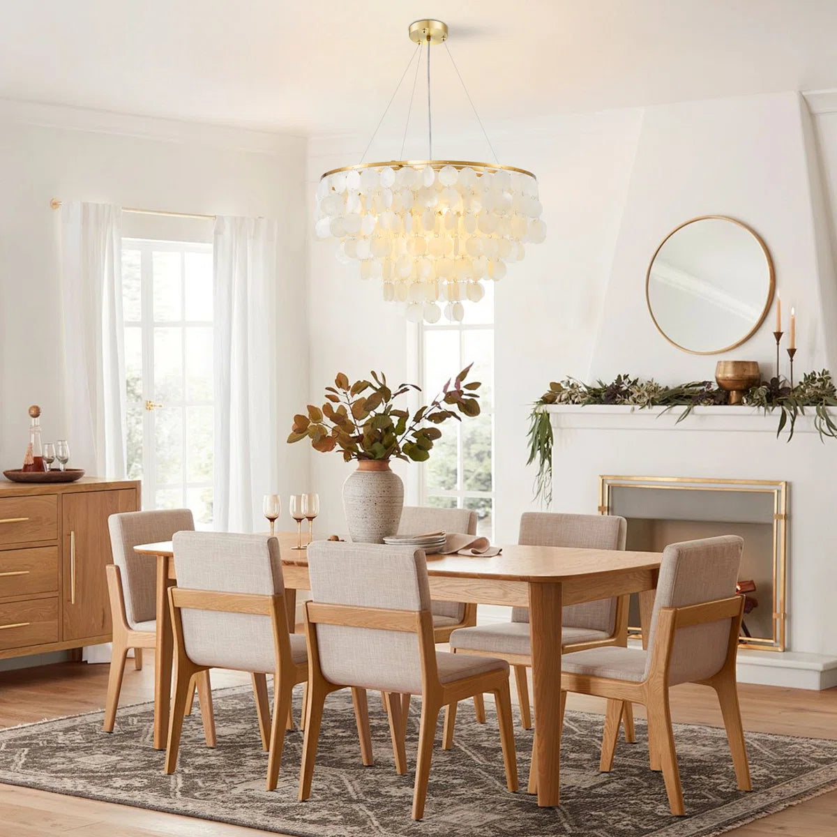 A modern dining room set featuring a light oak wood dining table and six upholstered chairs with light beige fabric, styled in a bright, contemporary dining room with a glass chandelier and fireplace.