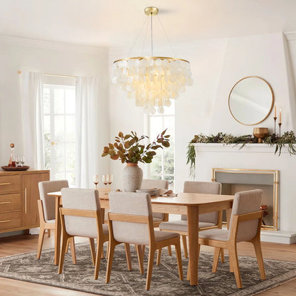 A modern dining room set featuring a light oak wood dining table and six upholstered chairs with light beige fabric, styled in a bright, contemporary dining room with a glass chandelier and fireplace.