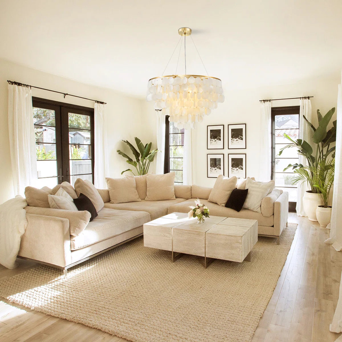 A modern beige sectional sofa in a bright, sunlit living room with a matching coffee table, textured rug, and a large glass chandelier.