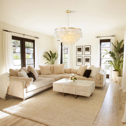 A modern beige sectional sofa in a bright, sunlit living room with a matching coffee table, textured rug, and a large glass chandelier.
