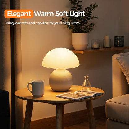 A mushroom-shaped table lamp with a glowing warm white shade and a beige base sits on a round wooden side table in a cozy living room setting. The scene includes a mug, an open magazine, a small glass bottle, a shelf with candles and a plant, and a sofa in the background.