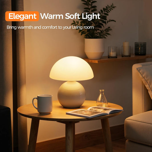 A mushroom-shaped table lamp with a glowing warm white shade and a beige base sits on a round wooden side table in a cozy living room setting. The scene includes a mug, an open magazine, a small glass bottle, a shelf with candles and a plant, and a sofa in the background.