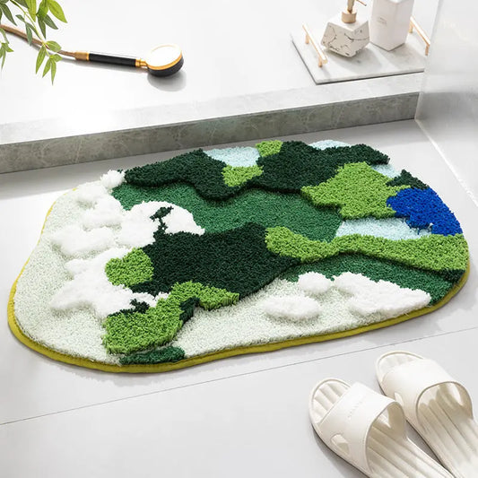 A rectangular, tufted bathroom mat with a moss green and blue pattern, placed on a light surface, with a pair of white sandals and a plant in the background.