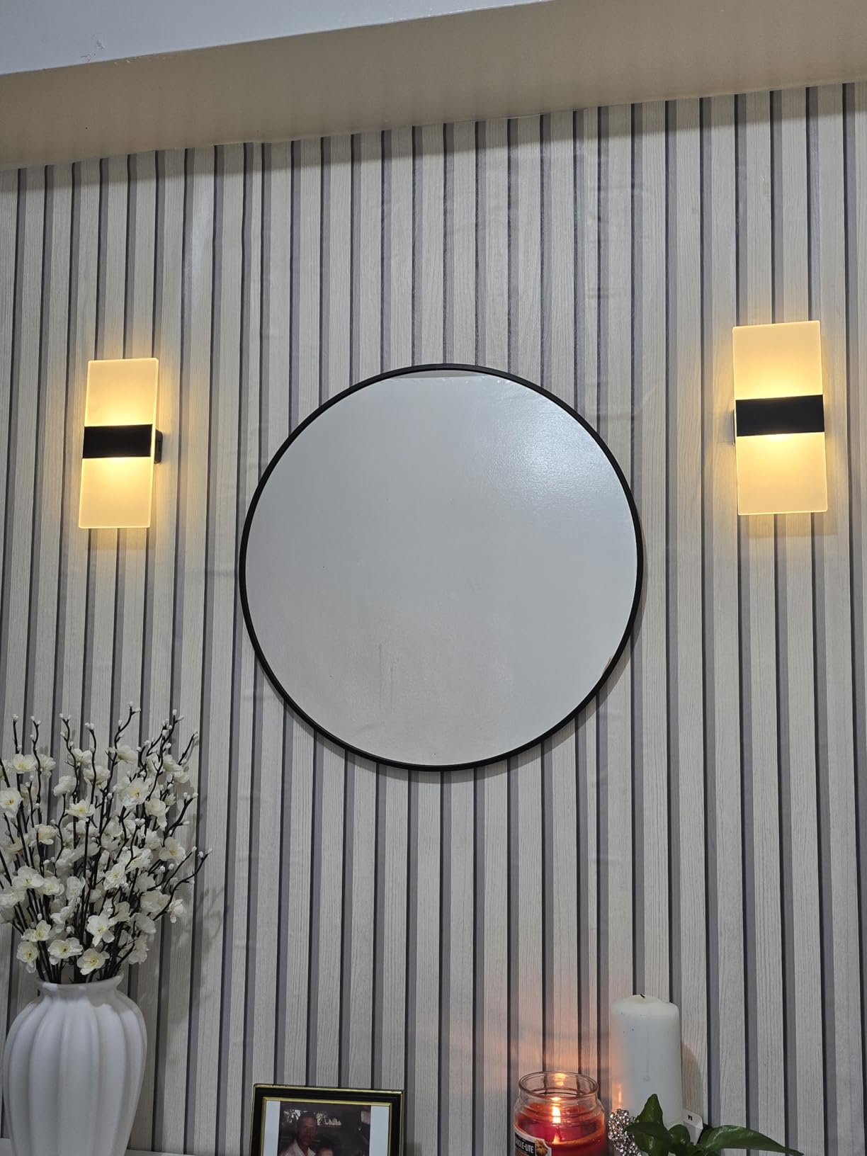 Round mirror on a wall with two wall lights and decorative items below.