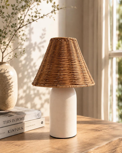 A table lamp with a natural woven rattan shade and a matte off-white cylindrical base, styled on a wooden table next to a vase and books in a warm, sunlit room.