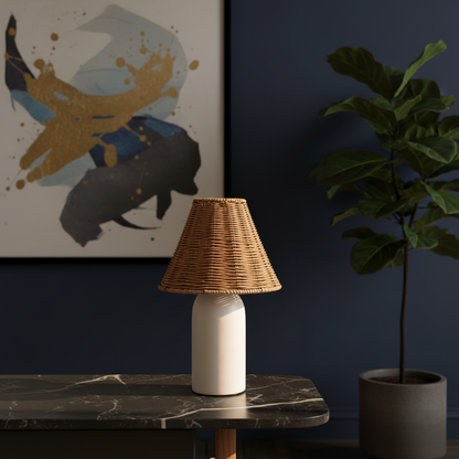 A table lamp with a natural woven rattan shade and a matte white cylindrical base sits on a dark marble-topped table in a styled room. A dark blue wall, an abstract fish painting, and a potted fiddle-leaf fig plant are visible in the background.