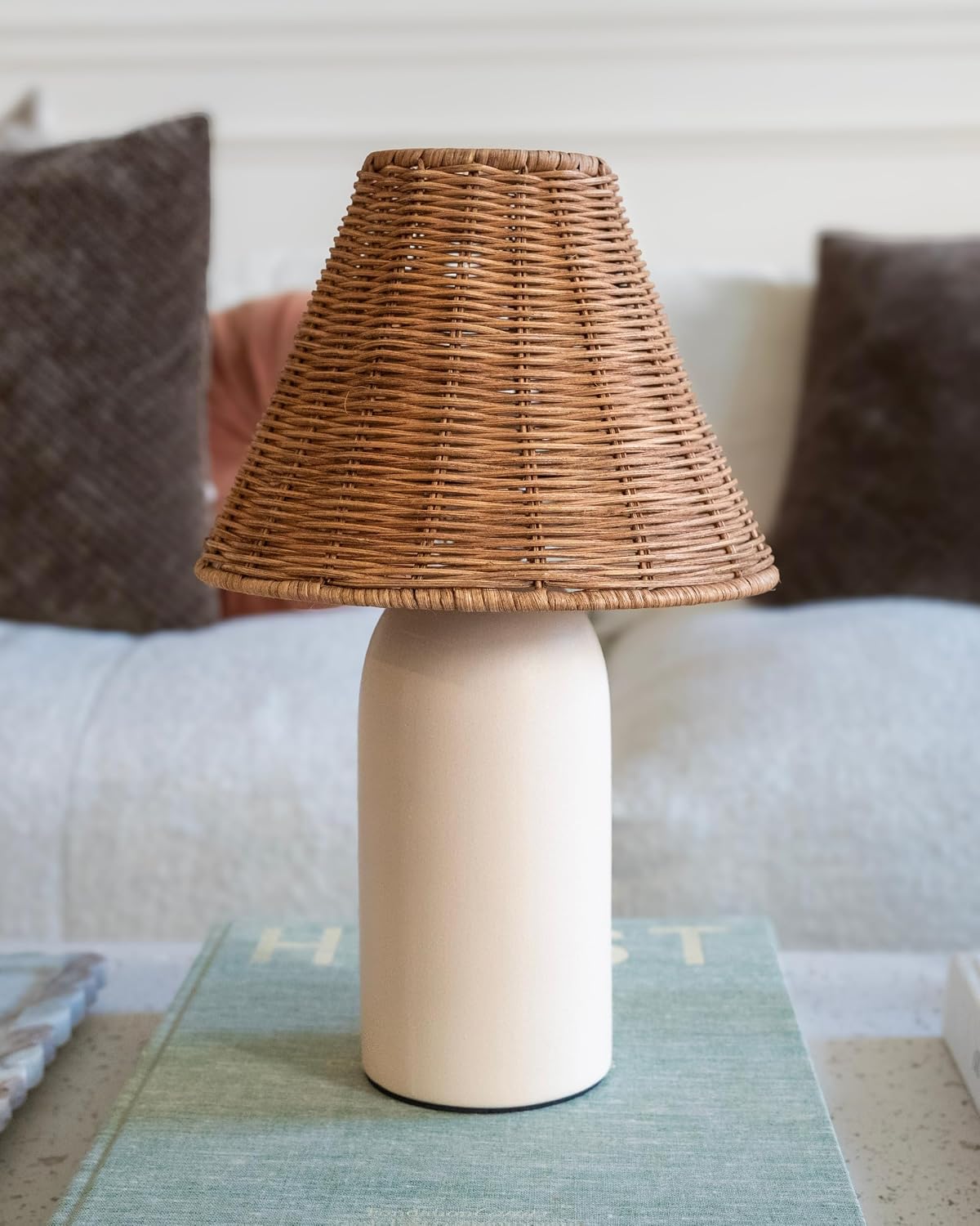 A table lamp with a natural woven wicker shade and a matte cream cylindrical base, displayed on a light blue-green textured surface in a softly blurred living room setting.