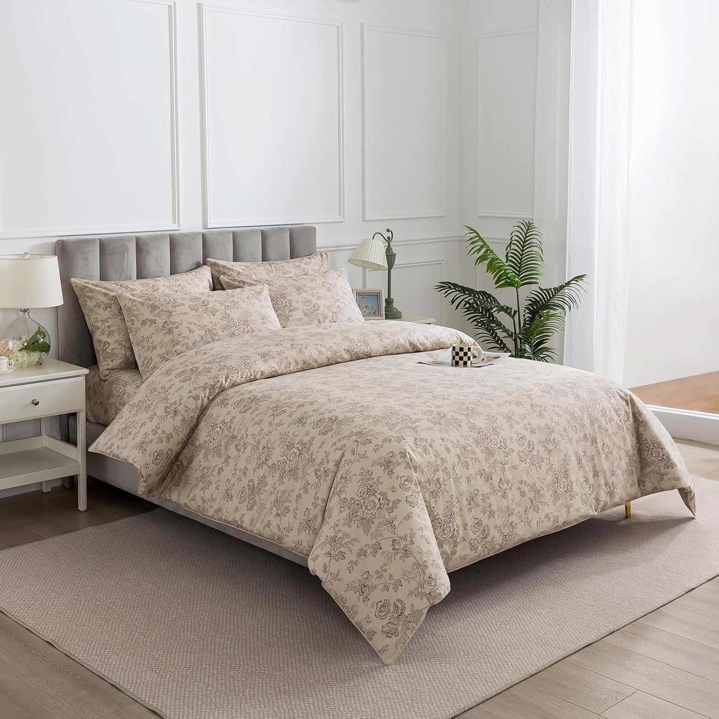 A beige and brown floral patterned duvet cover set with matching pillowcases on a gray upholstered bed, styled in a bright, modern bedroom with white walls, a bedside table, and a potted plant.