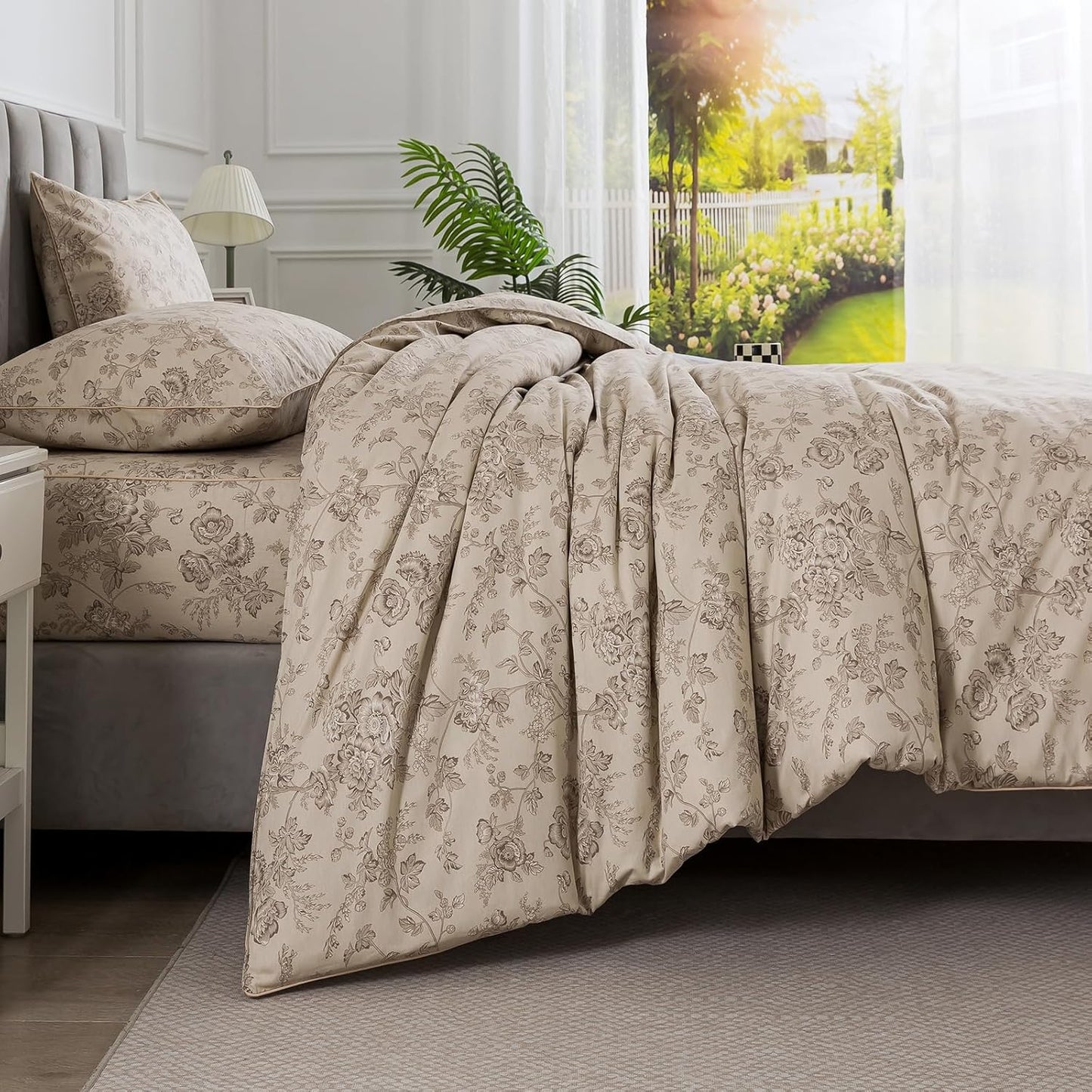 A beige and gray floral patterned comforter set styled on a bed in a bright, modern bedroom with white walls, a window, and a plant.