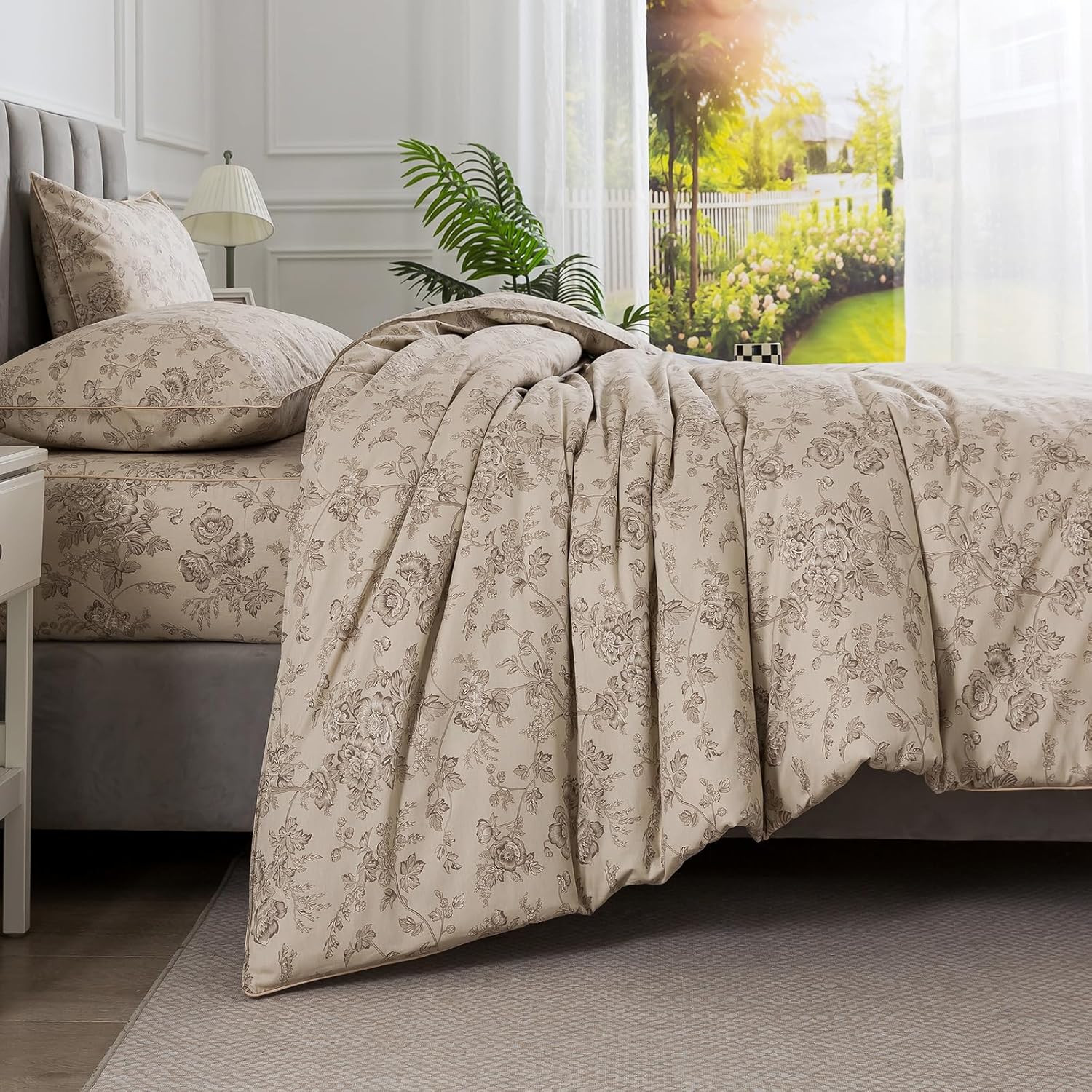 A beige and gray floral patterned comforter set styled on a bed in a bright, modern bedroom with white walls, a window, and a plant.