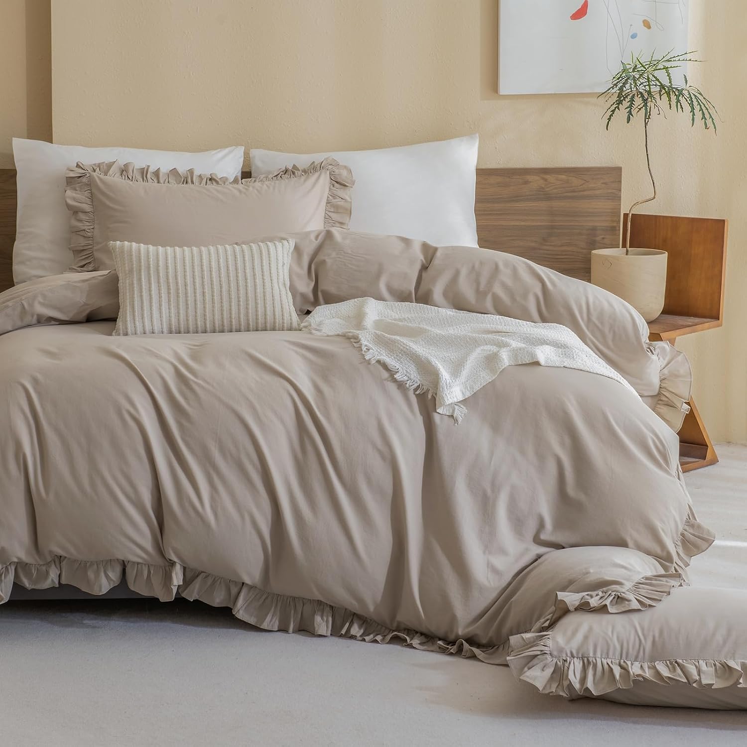A beige and white bedding set styled on a bed with a wooden headboard, featuring ruffled edges, striped pillows, and a white throw blanket in a warm, neutral bedroom setting.