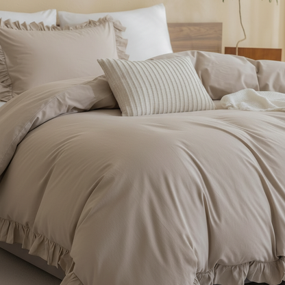 A beige ruffled duvet cover and pillow set styled on a bed in a softly lit bedroom, with a striped accent pillow and neutral walls in the background.