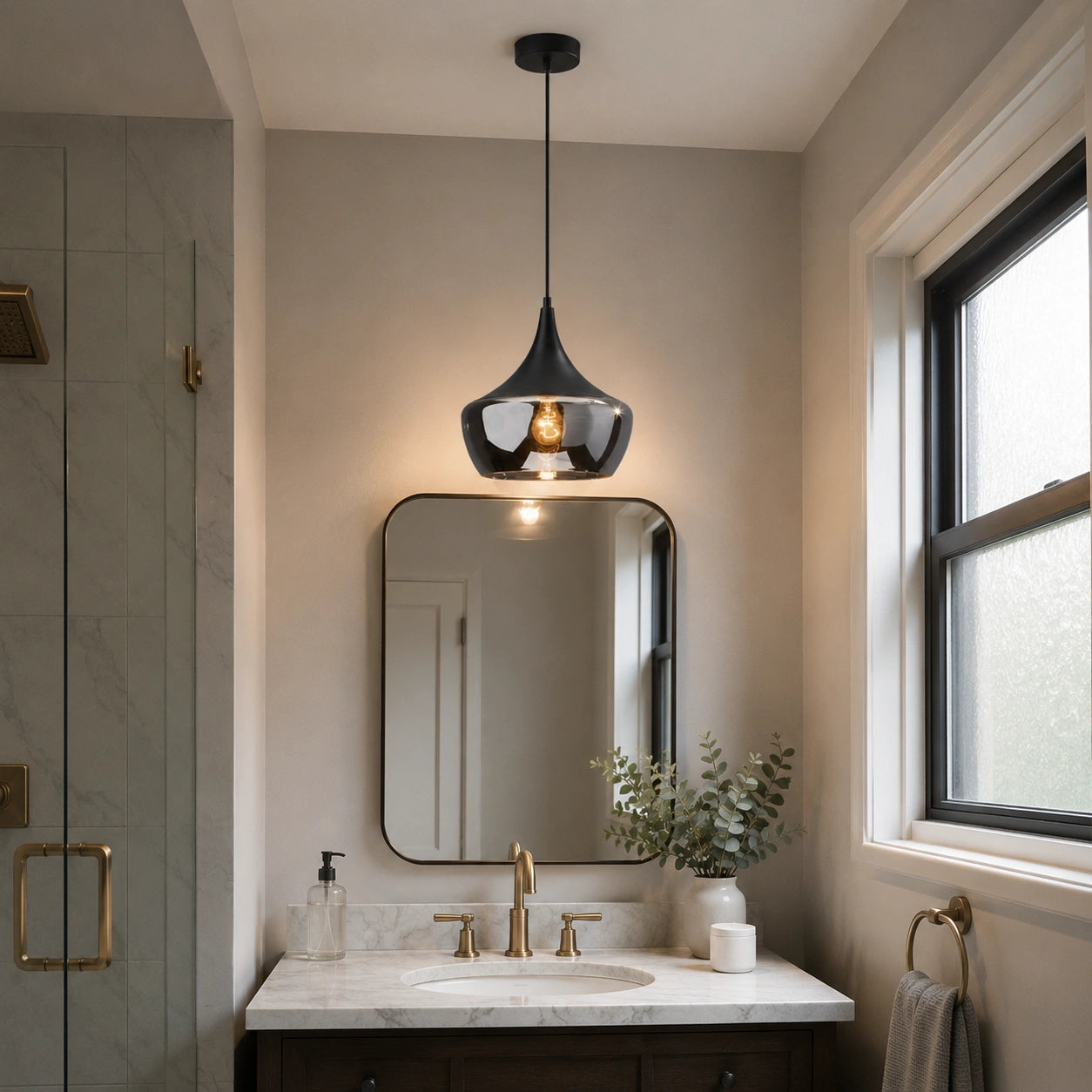 A modern black pendant light with a smoky glass shade hangs over a bathroom vanity with a white marble countertop, gold faucet, and a wall mirror.