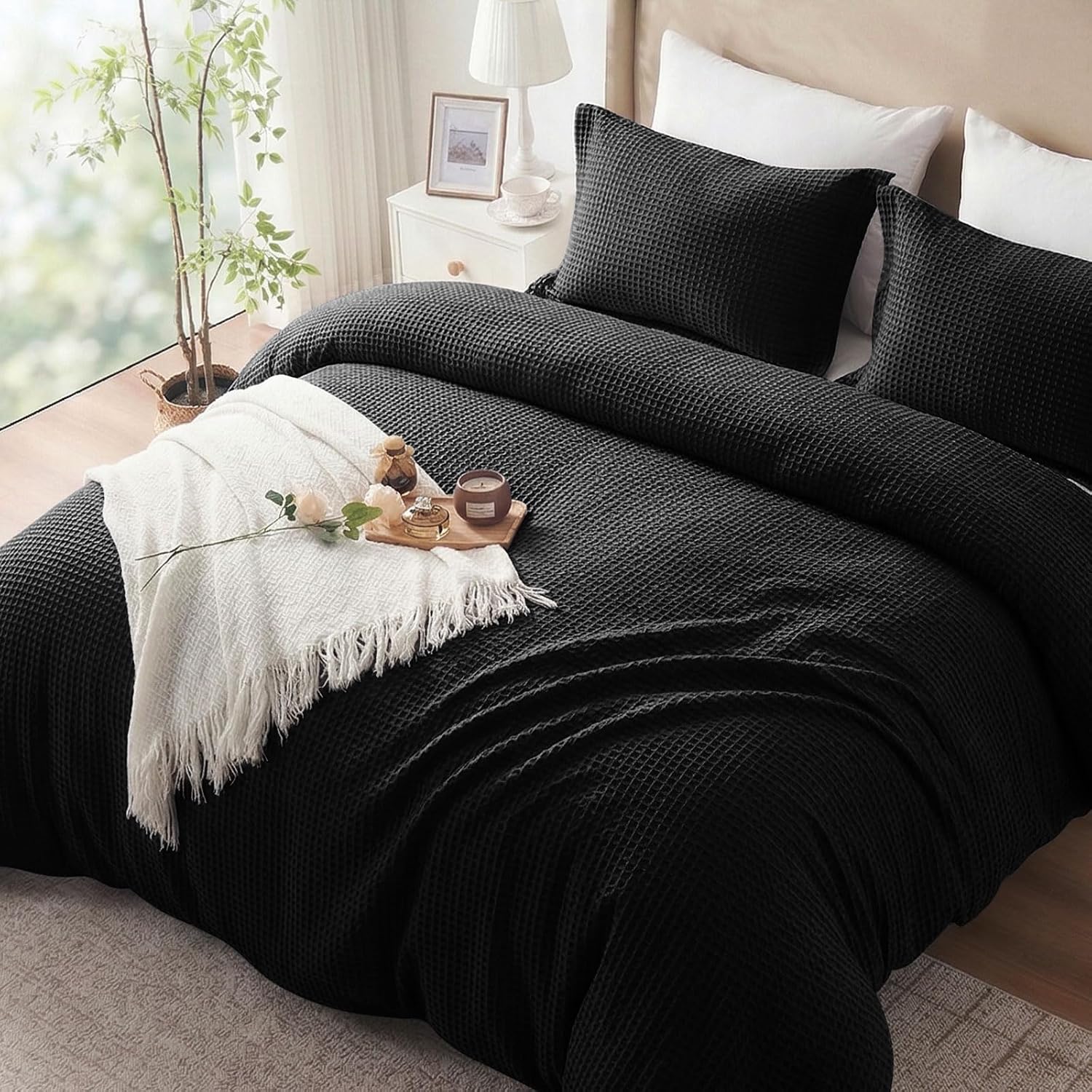 A black textured waffle-weave bedding set, including a duvet cover and two matching pillowcases, styled on a bed in a bright bedroom with a white throw blanket, pillows, and bedside table.