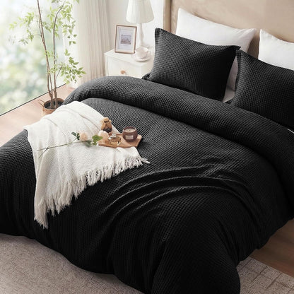 A black textured waffle-weave bedding set, including a duvet cover and two matching pillowcases, styled on a bed in a bright bedroom with a white throw blanket, pillows, and bedside table.