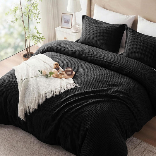 A black textured waffle-weave bedding set, including a duvet cover and two matching pillowcases, styled on a bed in a bright bedroom with a white throw blanket, pillows, and bedside table.