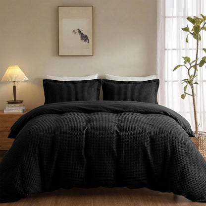 A black textured waffle-weave bedding set, including a duvet cover and two matching pillowcases, styled on a bed in a warm, modern bedroom setting.