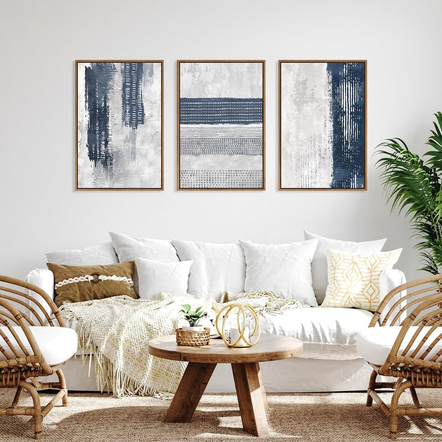 Three framed abstract wall art prints in navy, gray, and white hang above a white sofa in a bright living room with woven chairs, a round wooden coffee table, and a potted plant.