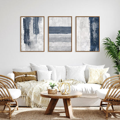 Three framed abstract wall art prints in navy, gray, and white hang above a white sofa in a bright living room with woven chairs, a round wooden coffee table, and a potted plant.