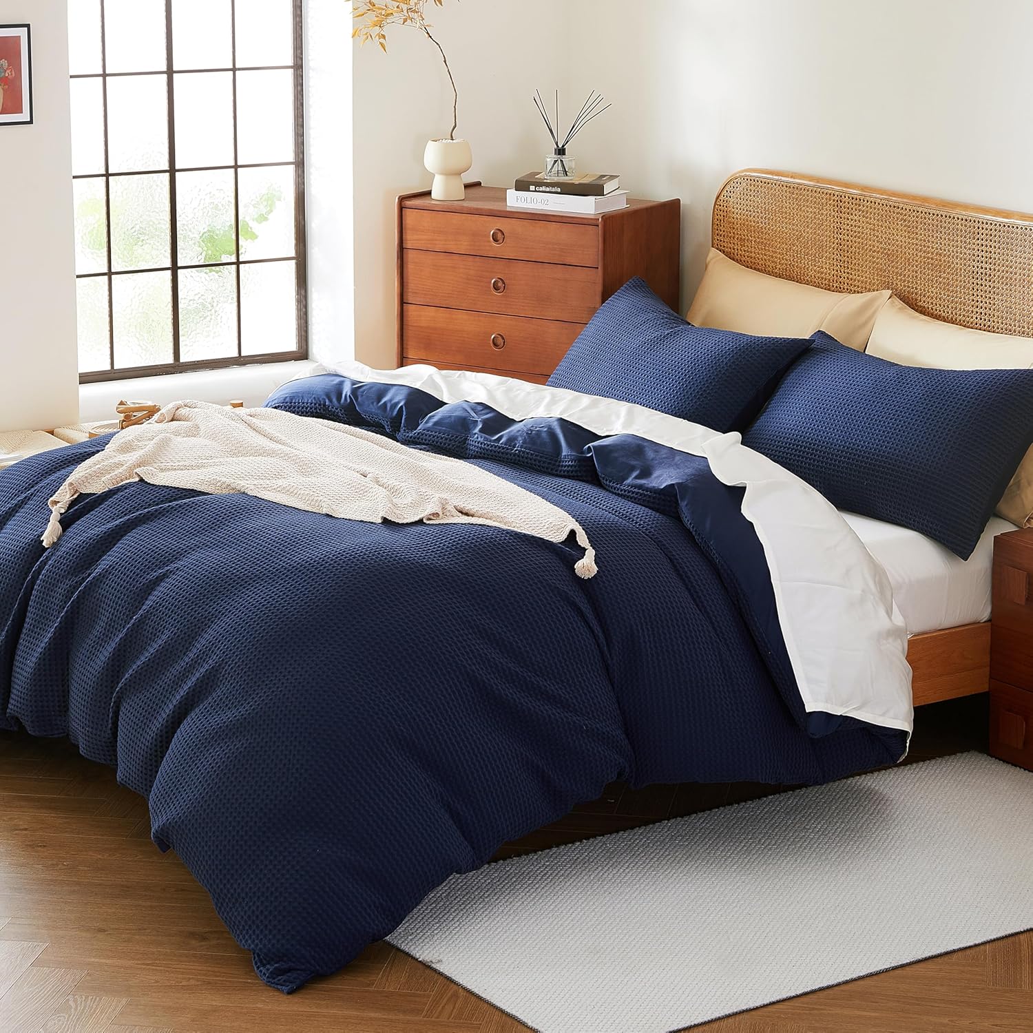 A bed dressed in a navy blue textured waffle-weave duvet cover and matching pillowcases, with a white sheet and cream throw blanket, shown in a styled bedroom setting.
