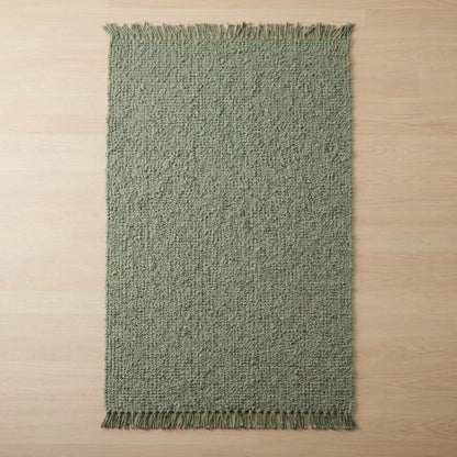 A rectangular woven rug in a muted sage green color with fringed edges, laid flat on a light wood floor.