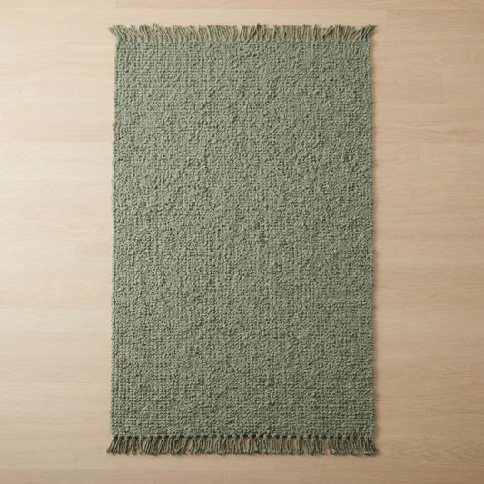 A rectangular woven rug in a muted sage green color with fringed edges, laid flat on a light wood floor.
