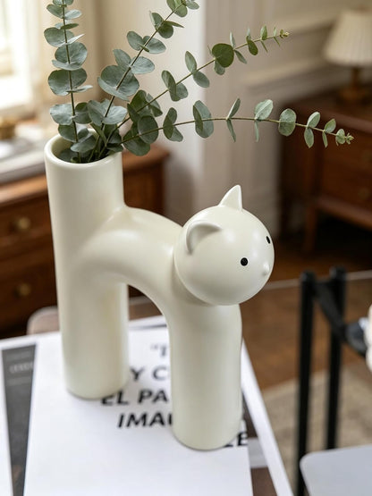 White ceramic vase shaped like a cat with green leaves on top, placed on a surface.