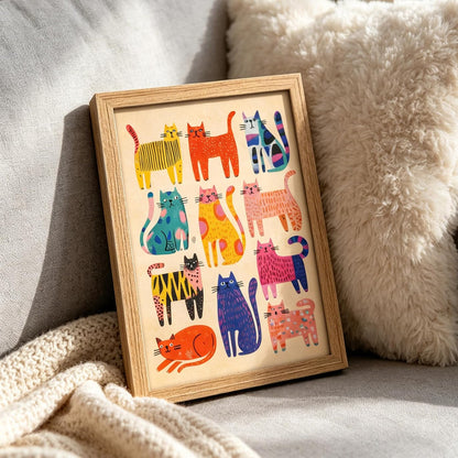 A framed art print featuring a colorful grid of whimsical cat illustrations in various patterns and colors, set in a light wood frame and styled on a cozy beige sofa next to a fluffy cream pillow.