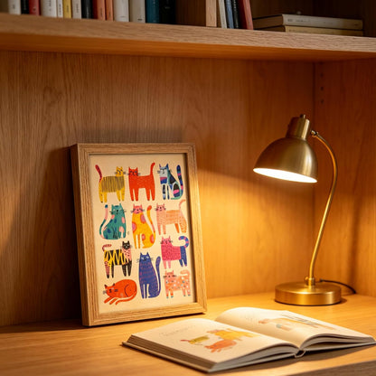 A framed art print featuring colorful illustrated cats, displayed on a wooden bookshelf next to a lit brass desk lamp and an open book.