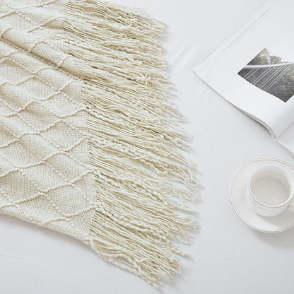Beige woven blanket with fringe on a white surface with a magazine and cup.