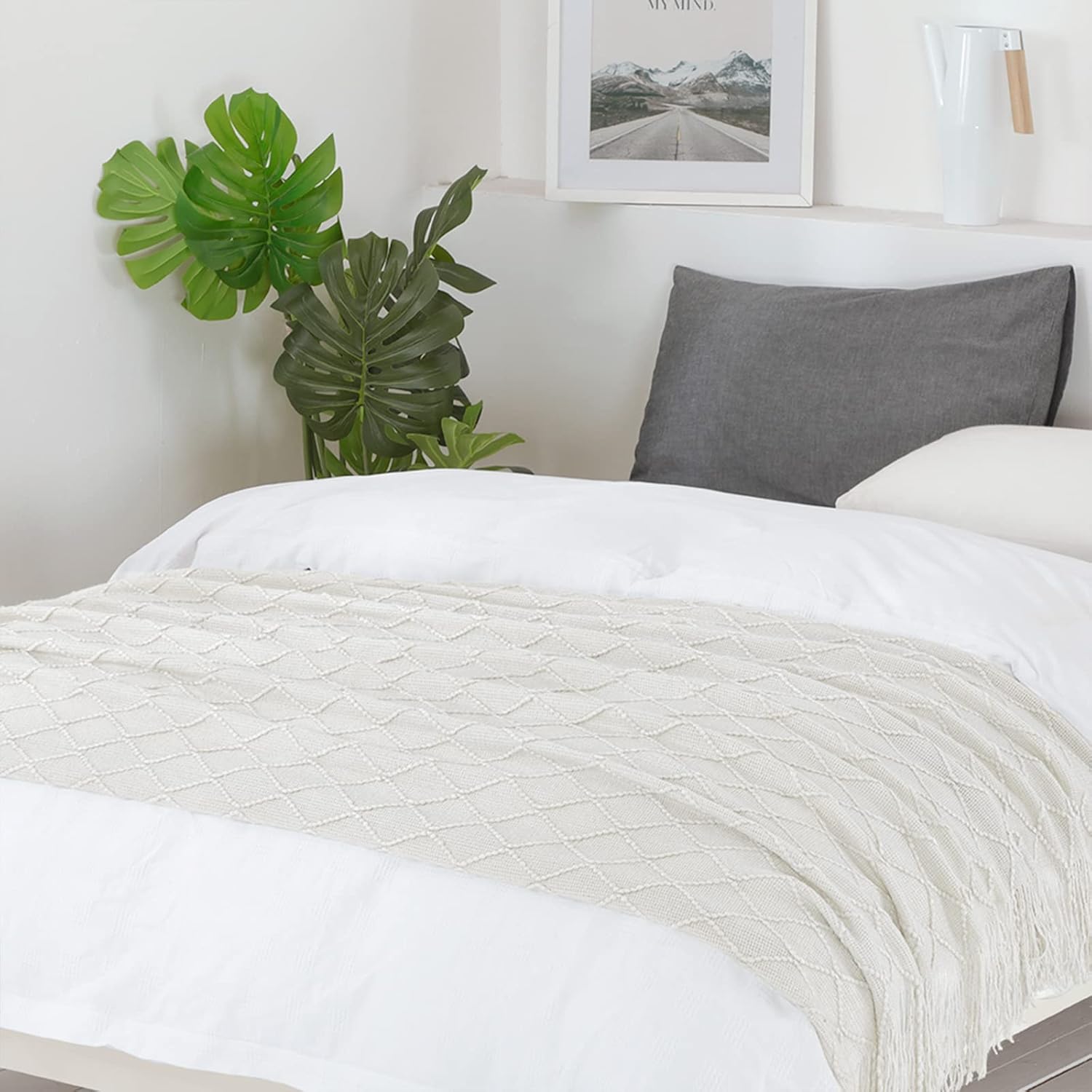 Bed with white bedding and a gray pillow, plant, and framed picture in the background.