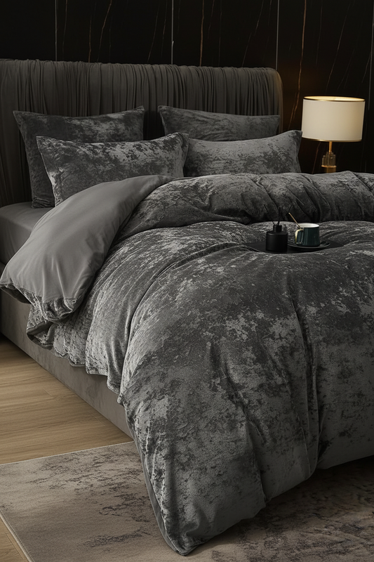A bed dressed in a dark gray crushed-velvet duvet cover and matching pillowcases, styled in a modern bedroom with a dark upholstered headboard, a lit bedside lamp, and a rug.