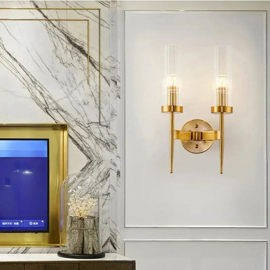 A modern double head gold wall sconce with clear glass lampshades, featuring a gold finish, mounted on a wall.