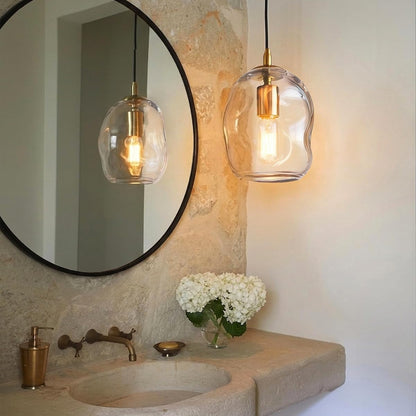 A modern pendant light with an organic, wavy clear glass shade and a brass-colored bulb holder hangs over a stone bathroom vanity next to a large round mirror.