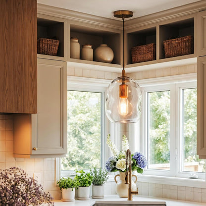 A modern kitchen corner with a clear glass globe pendant light hanging over a sink. The space features warm wood cabinetry, white subway tile backsplash, white countertops, and a window looking out to green trees.