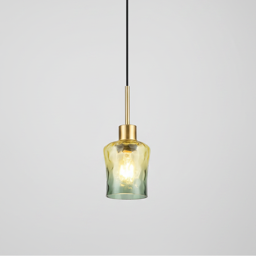 A modern pendant light with a textured, hammered glass shade that transitions from a warm yellow-gold at the top to a muted sage green at the bottom, suspended by a slim black cord and a brushed brass rod.