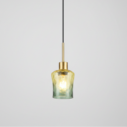 A modern pendant light with a textured, hammered glass shade that transitions from a warm yellow-gold at the top to a muted sage green at the bottom, suspended by a slim black cord and a brushed brass rod.