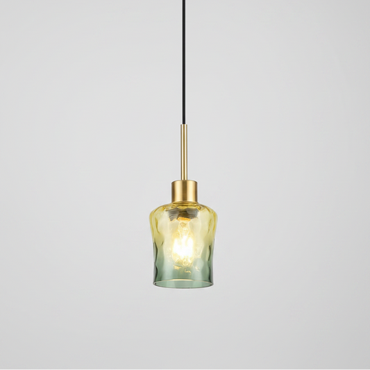 A modern pendant light with a textured, hammered glass shade that transitions from a warm yellow-gold at the top to a muted sage green at the bottom, suspended by a slim black cord and a brushed brass rod.