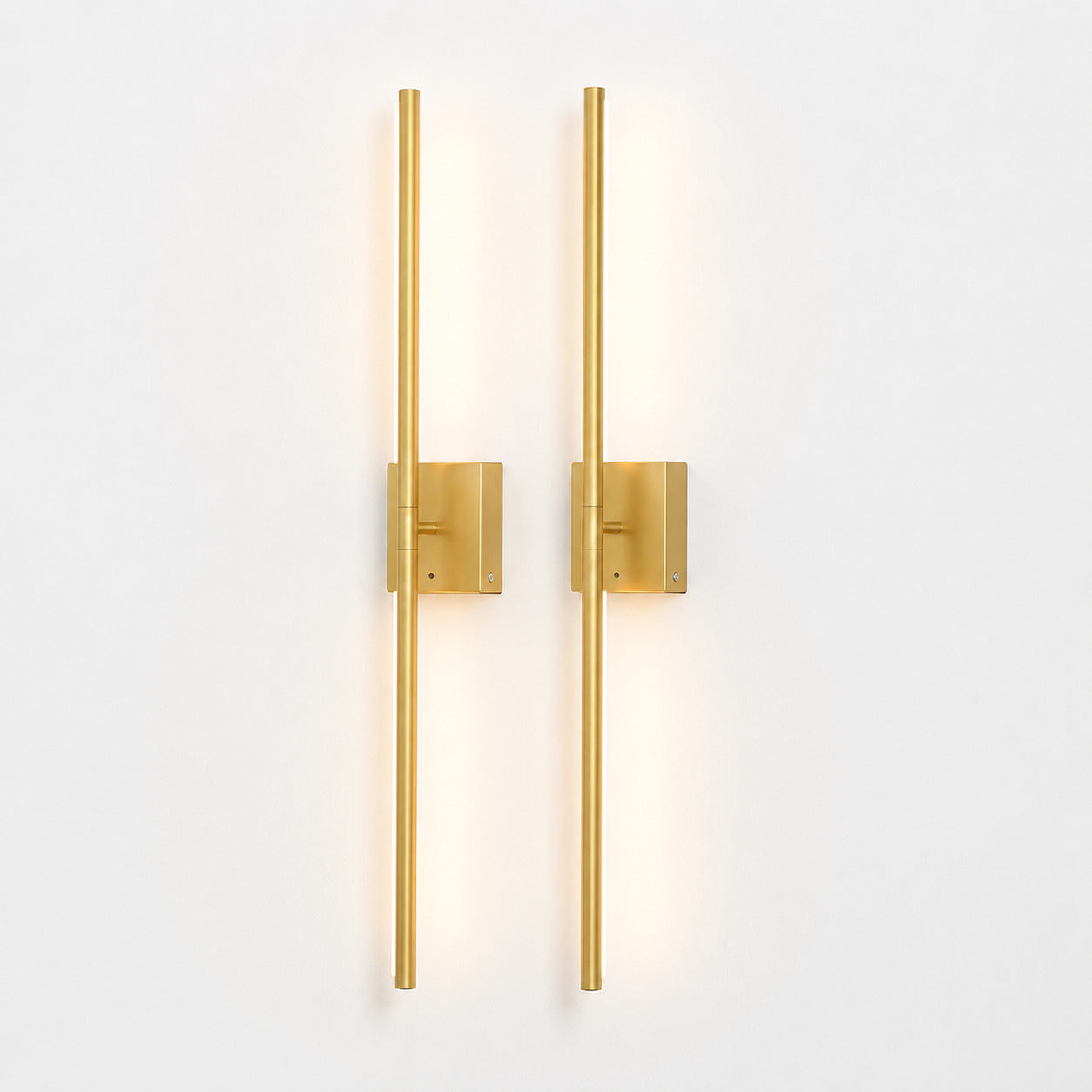 Two modern vertical wall sconces with slim cylindrical rods and square backplates in a brushed gold finish, mounted side by side on a plain white wall and casting a warm light.