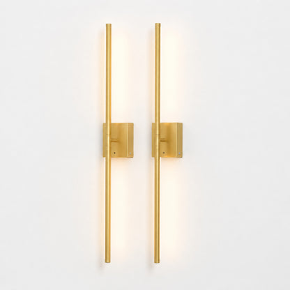 Two modern vertical wall sconces with slim cylindrical rods and square backplates in a brushed gold finish, mounted side by side on a plain white wall and casting a warm light.